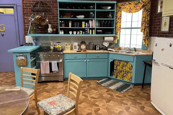 Le décor reconstitué de la cuisine de Monica dans Friends : meubles turquoise, mur de brique, plan de travail en L sous la fenêtre.
